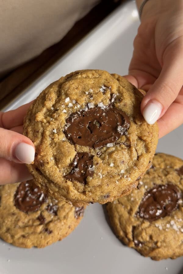 A person holds a chocolate chip cookie topped with sea salt flakes, with two more cookies visible below on a white plate.