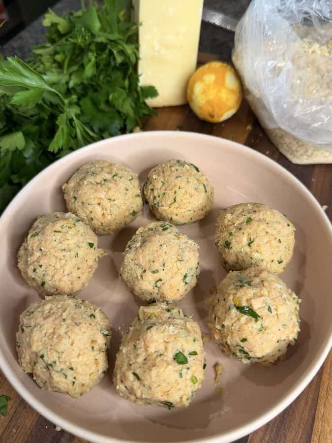 Seven raw salmon meatballs in a round bowl, with parsley, a block of cheese, a lemon, and a bag of breadcrumbs in the background.