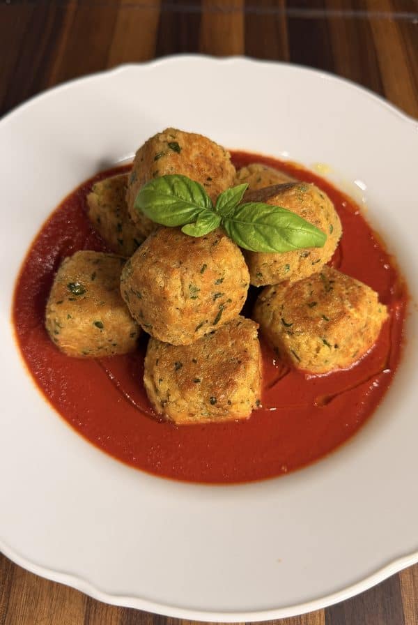 A pile of golden-brown herb meatballs in a pool of red tomato sauce, garnished with a fresh basil leaf on a white plate.