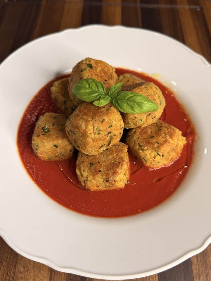 A pile of golden-brown herb meatballs in a pool of red tomato sauce, garnished with a fresh basil leaf on a white plate.