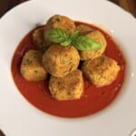 A pile of fried vegetable fritters in a red tomato sauce, garnished with a fresh basil leaf on a white plate.