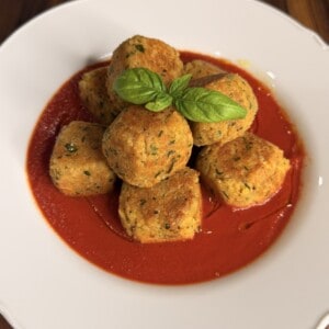 A pile of fried vegetable fritters in a red tomato sauce, garnished with a fresh basil leaf on a white plate.