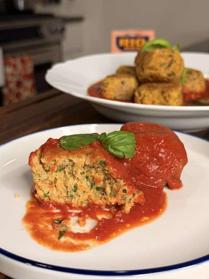 A meatball cut in half, topped with tomato sauce and a basil leaf, on a white plate with more meatballs in the background.