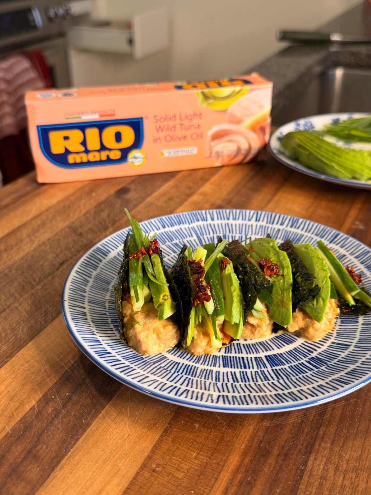Four tuna and avocado nori tacos topped with scallions and chili flakes on a blue and white patterned plate, with a box of Rio Mare tuna in the background.