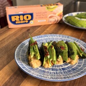 Four tuna and avocado nori tacos topped with scallions and chili flakes on a blue and white patterned plate, with a box of Rio Mare tuna in the background.