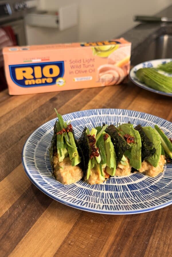 Four tuna and avocado nori tacos topped with scallions and chili flakes on a blue and white patterned plate, with a box of Rio Mare tuna in the background.