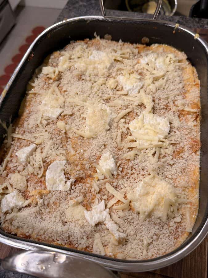 A rectangular baking dish filled with an unbaked pasta dish topped with shredded cheese and dollops of soft cheese.