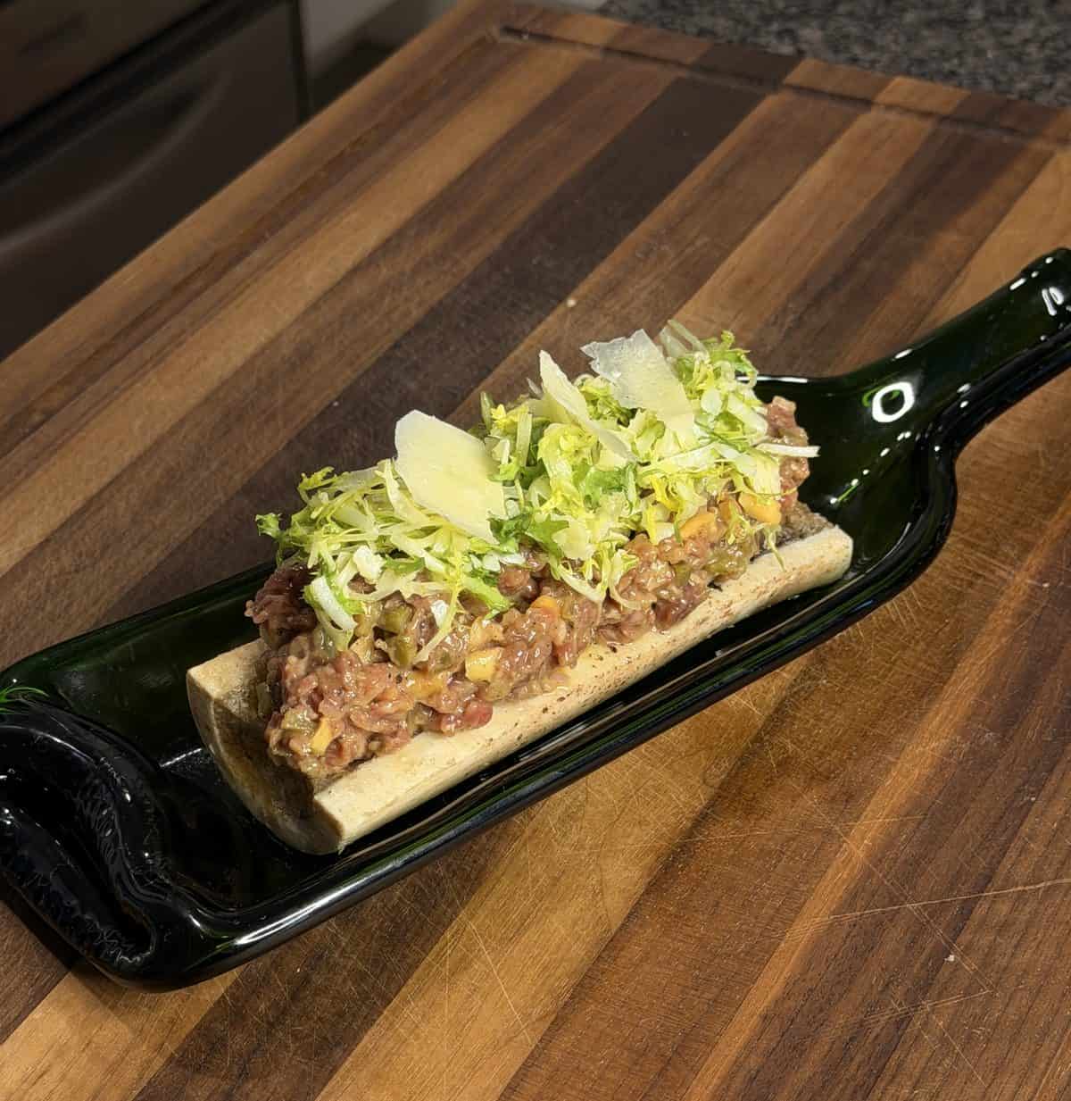 Beef tartare served on a roasted bone marrow in a repurposed glass bottle tray, topped with shaved cheese and greens.