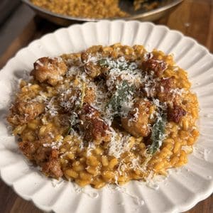 A plate of creamy risotto topped with sausage pieces, fried sage leaves, and grated cheese, with a pan of more risotto in the background on a wooden counter.
