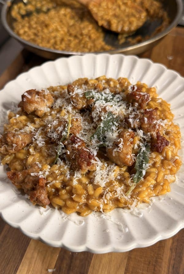 A plate of creamy risotto topped with sausage pieces, fried sage leaves, and grated cheese, with a pan of more risotto in the background on a wooden counter.