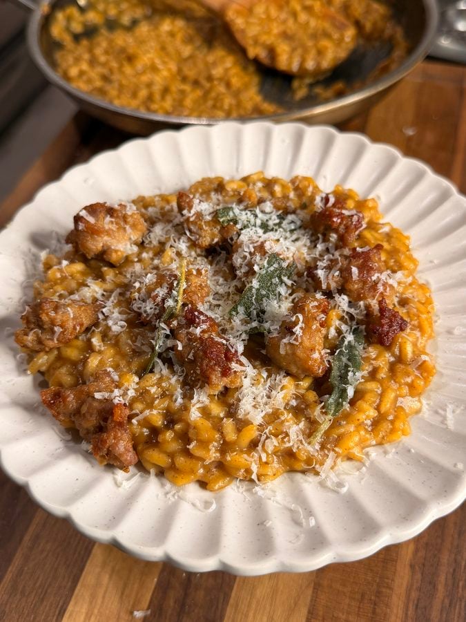 A plate of creamy risotto topped with sausage pieces, fried sage leaves, and grated cheese, with a pan of more risotto in the background on a wooden counter.