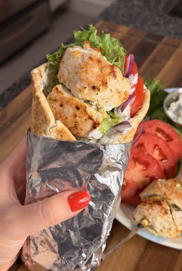 A hand holding a chicken souvlaki wrap in foil, with side portions of tomato slices, chicken skewers, and tzatziki sauce on a plate.