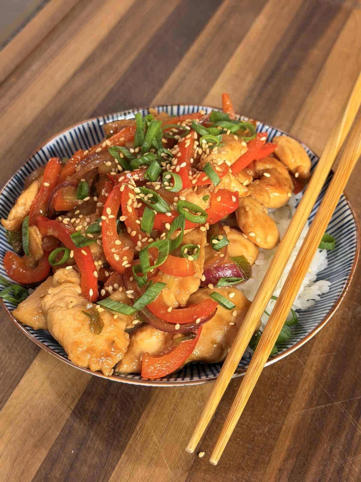 A bowl of chicken and pepper stir-fry topped with sesame seeds and green onions, served over white rice with a pair of wooden chopsticks.