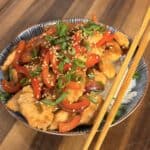 A bowl of chicken and pepper stir-fry topped with sesame seeds and green onions, served over white rice with a pair of wooden chopsticks.