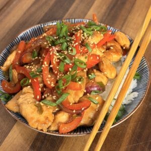 A bowl of chicken and pepper stir-fry topped with sesame seeds and green onions, served over white rice with a pair of wooden chopsticks.