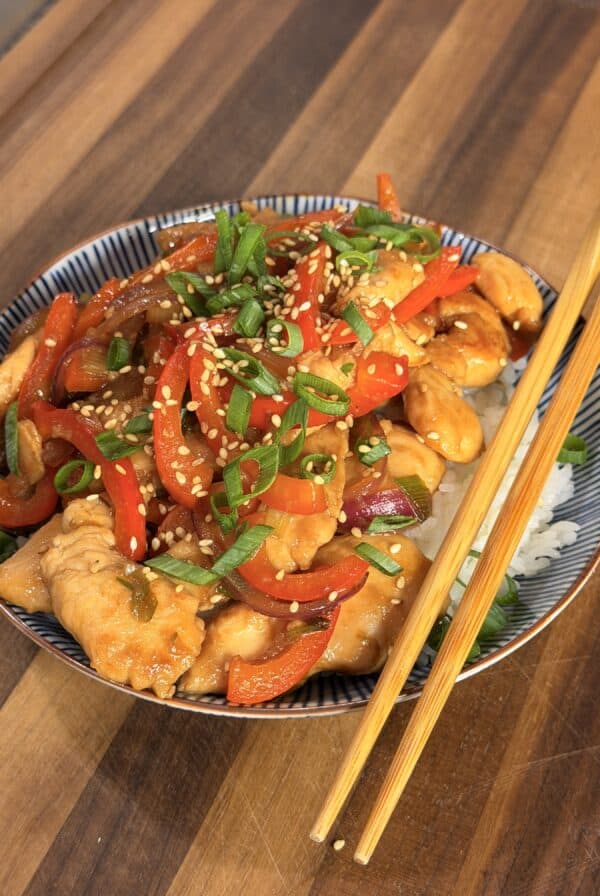 A bowl of chicken and pepper stir-fry topped with sesame seeds and green onions, served over white rice with a pair of wooden chopsticks.