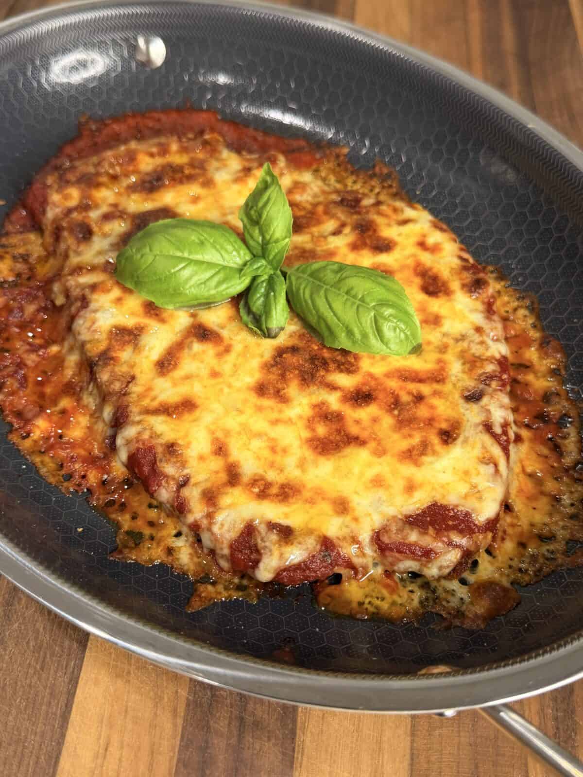 Chicken parmesan topped with melted cheese and fresh basil leaves in a nonstick skillet.