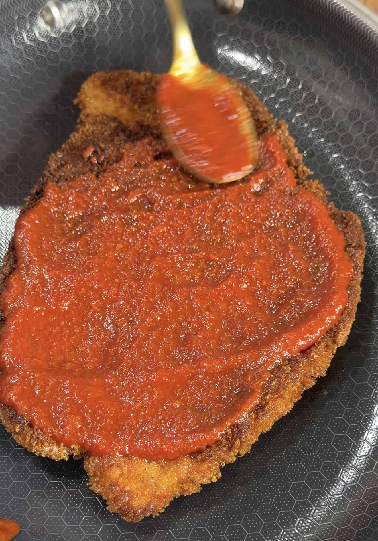 A metal spoon spreads red tomato sauce over a breaded, fried cutlet in a skillet.