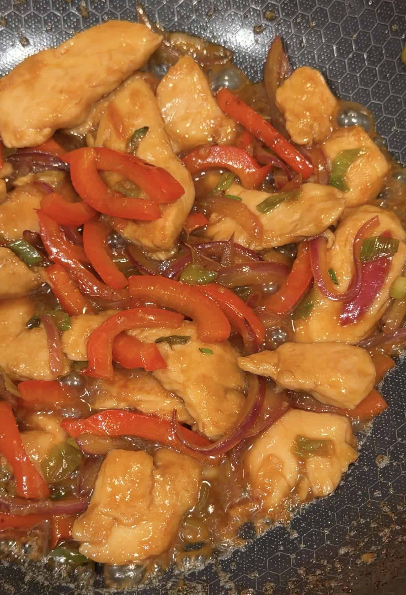 Stir-fry with chicken pieces, sliced red bell peppers, and red onion in a glazed sauce, cooking in a hexagonal-patterned skillet.
