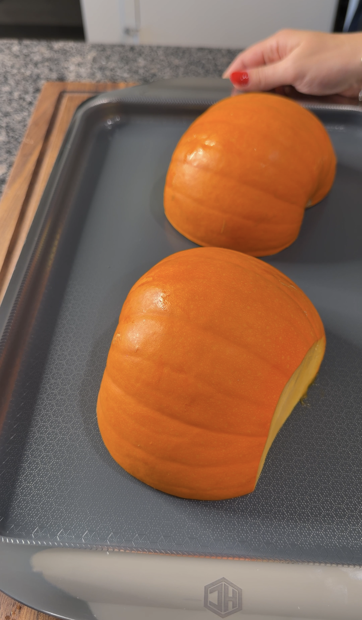 Two orange pumpkin halves are placed cut side down on a gray baking sheet, ready for roasting. A hand with red nail polish rests in the background near the corner of the pan.