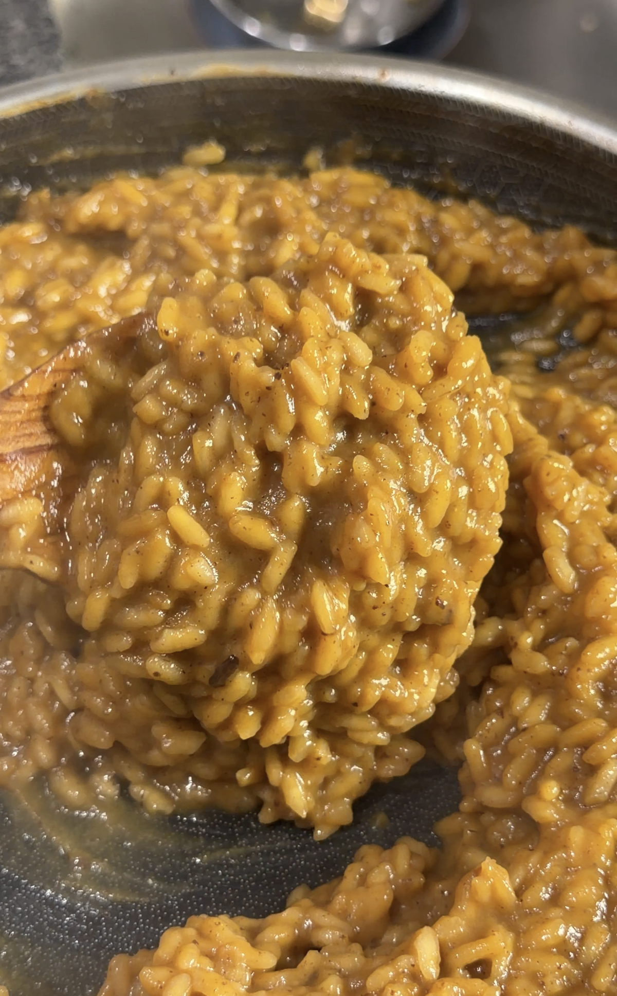 Close-up of creamy brown risotto being stirred with a wooden spoon in a pan. The grains of rice are well-cooked and coated in a thick, rich-looking sauce.