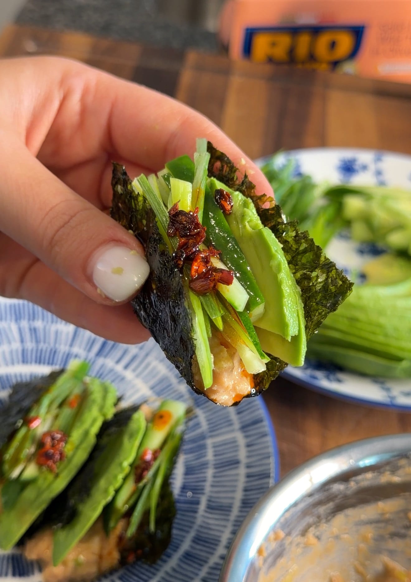 A hand holds a nori wrap filled with salmon, sliced avocado, and cucumber, topped with chili crisp.