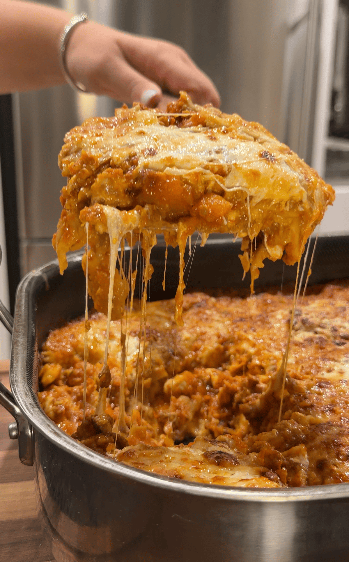 A serving spoon lifting a large, cheesy slice of lasagna from a pan, with strings of melted mozzarella stretching down toward the rest of the dish.