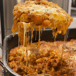 A serving spoon lifting a large, cheesy slice of lasagna from a pan, with strings of melted mozzarella stretching down toward the rest of the dish.