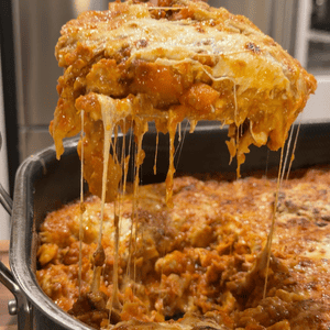 A serving spoon lifting a large, cheesy slice of lasagna from a pan, with strings of melted mozzarella stretching down toward the rest of the dish.