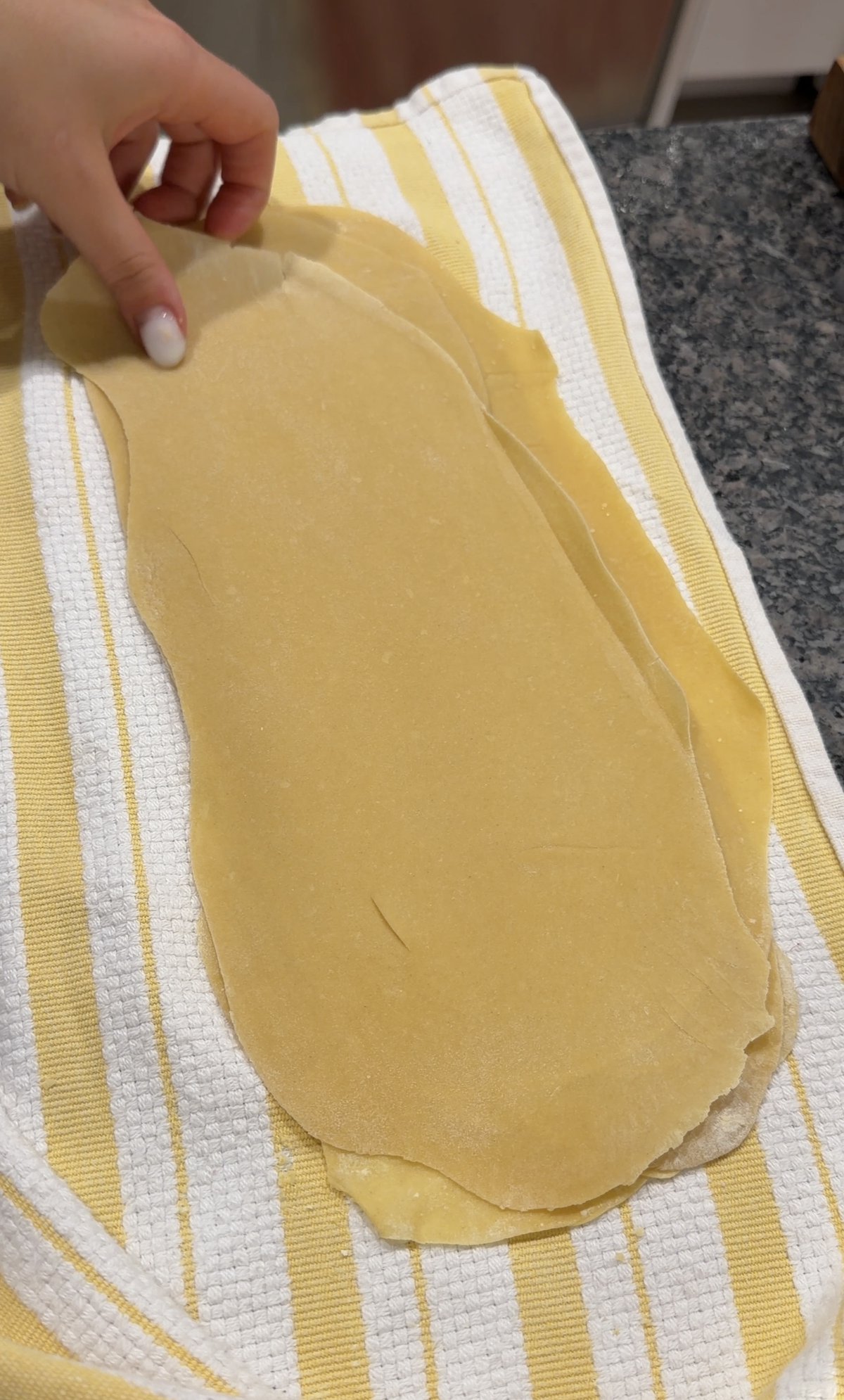 A hand places a thin sheet of fresh pasta dough onto a yellow and white striped kitchen towel.
