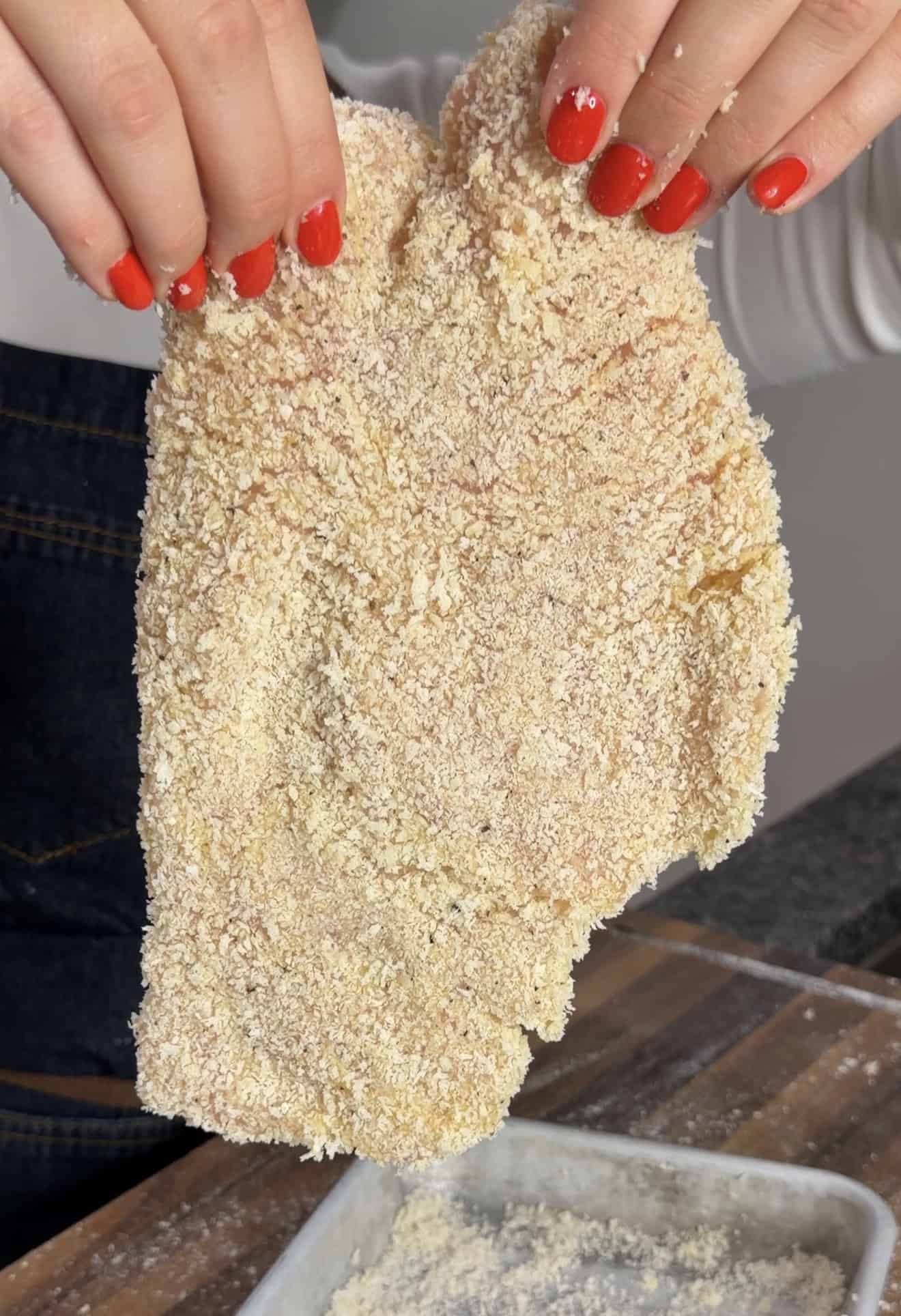Hands with red fingernails holding a large, breaded raw chicken cutlet.