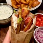 A hand holding a pita wrap filled with seasoned chicken, lettuce, tomatoes, onion, fries, and sauce. Bowls of tzatziki, chicken, chopped tomatoes, and sliced onions are in the background on a wooden table.